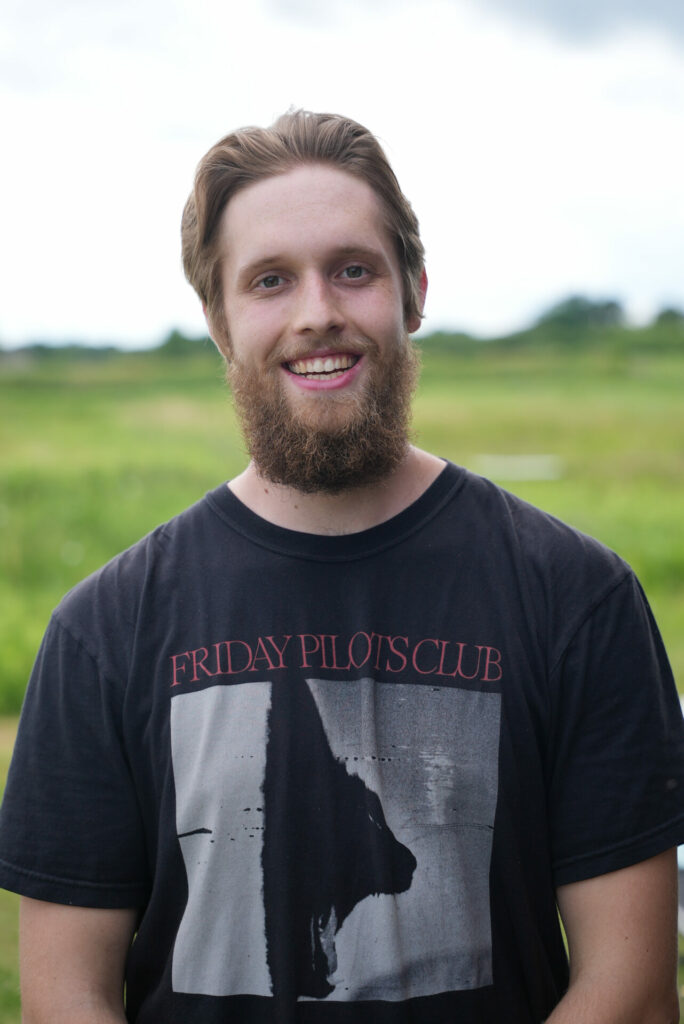 aquaculture intern, Wyatt Slack, stands in a black t-shirt in front of a green field