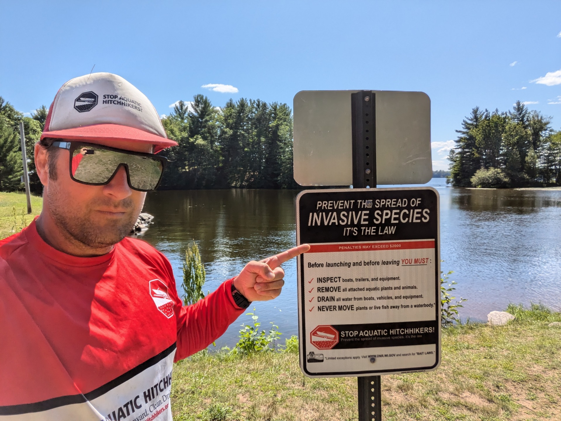 A man in reflective sunglasses wearing a Stop Aquatic Hitchikers shirt points to a sign telling boaters to remove invasive species from their boats