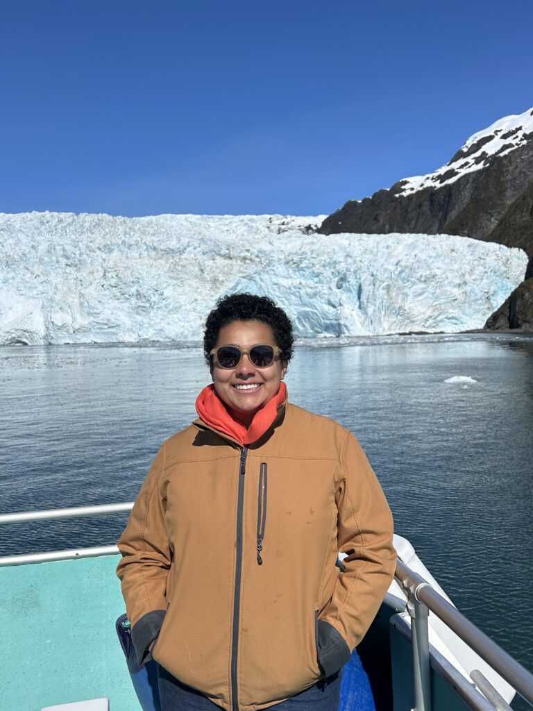 Marian Azeem Angel in a brown jacket stands on a boat overlooking the Holgate Glacier in Alaska
