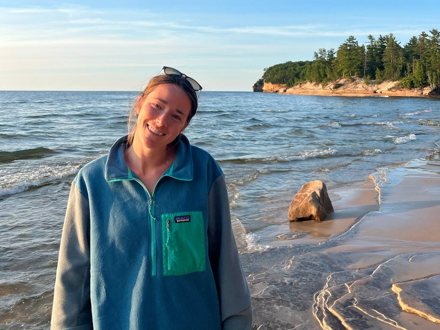 Cailin Young wears a blue jacket and stands on the shores of Lake Superior, with a tree-lined bluff in the background.