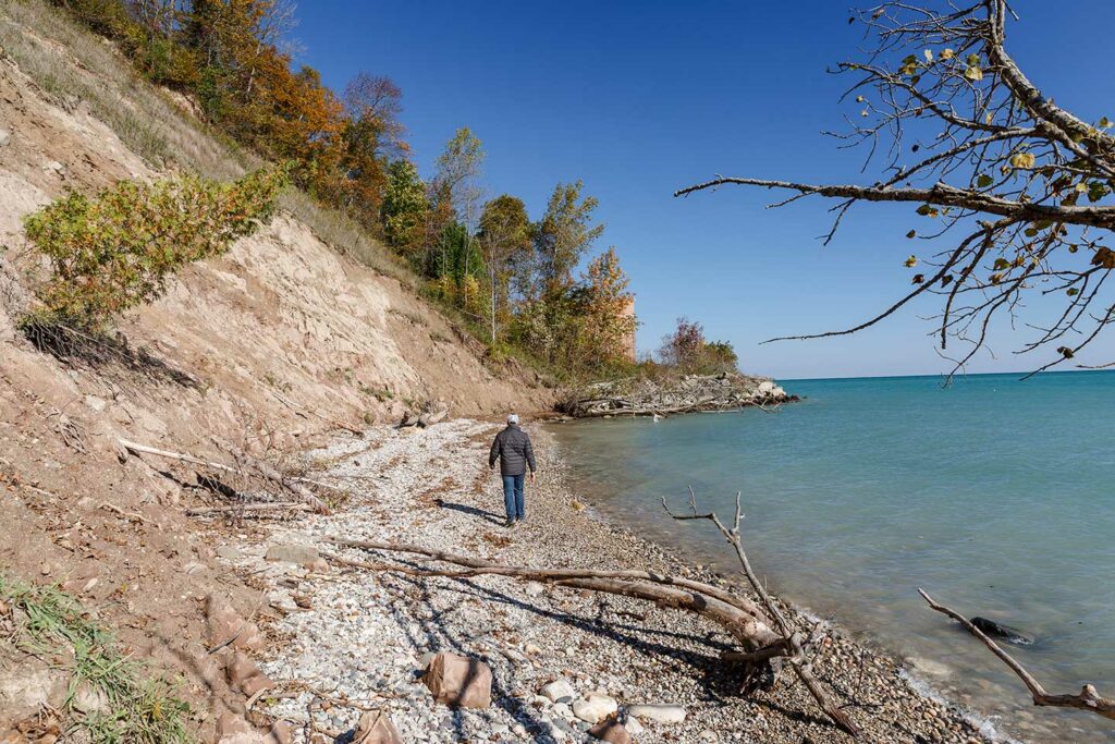 A man in a black coat walks along a Lake Michigan beach at the bottom of a sandy, eroding bluff