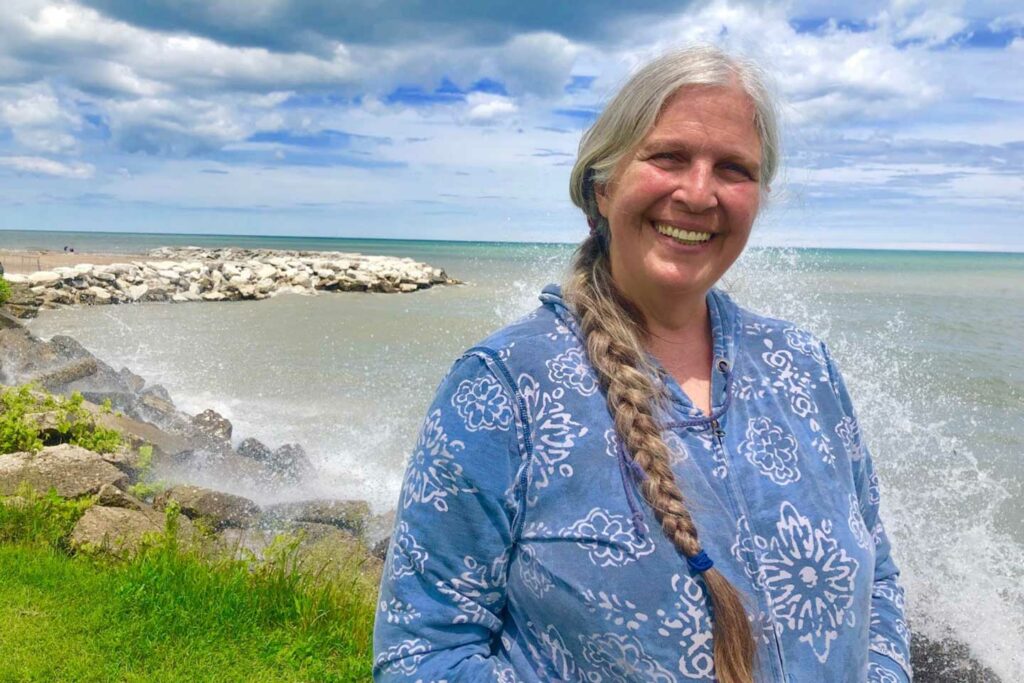 A woman with a long white braid wearing a blue patterend shirt smiles on the shores of Lake Michigan