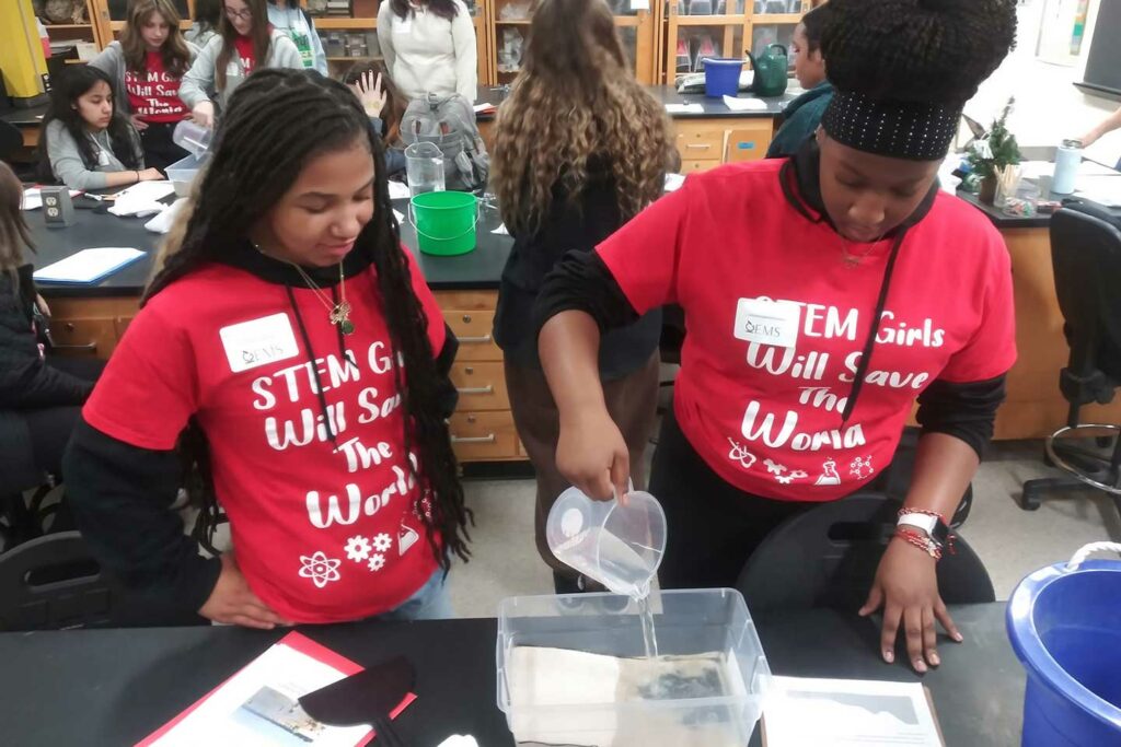 Two students in red t-shirts that say "STEM girls will save the world" pour water into a sand-filled plastic container to prepare for the wave tank activity.