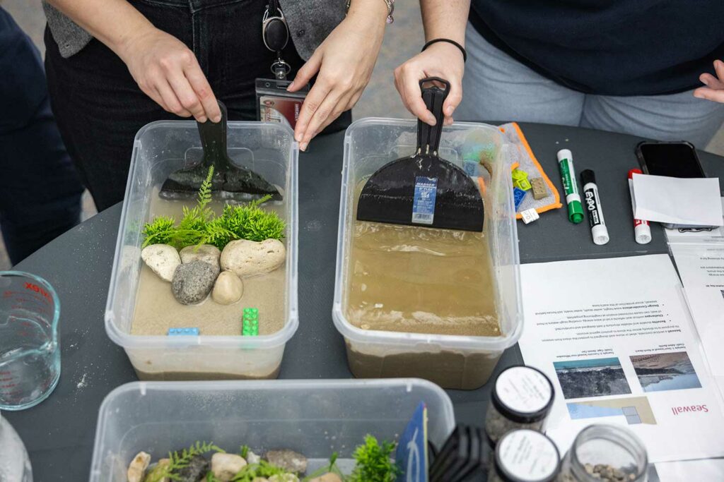 Students experiment with two wave tanks. The tanks are shoebox-sized plastic containers filled with sand and water. One contains rocks and plastic vegetation and the other does not.