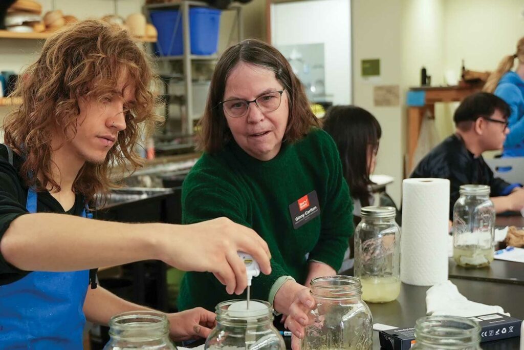 In the foreground, one person lifts a lid from a glass jar and another person reaches for a second glass jar. In the background, people read papers and look at objects on desks.