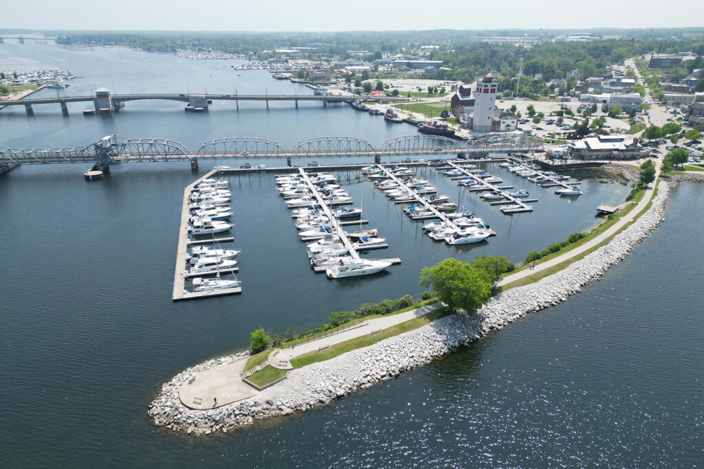 Aerial view of a marina filled with rows of docked white boats along a calm waterway, bordered by a curved rock-lined shoreline with a walking path, trees, and park benches in the foreground. A metal truss bridge and a lift bridge span the water toward a small waterfront town with buildings and parking areas in the background.