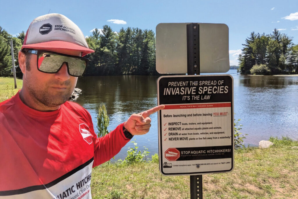 A man in reflective sunglasses wearing a Stop Aquatic Hitchikers shirt points to a sign telling boaters to remove invasive species from their boats